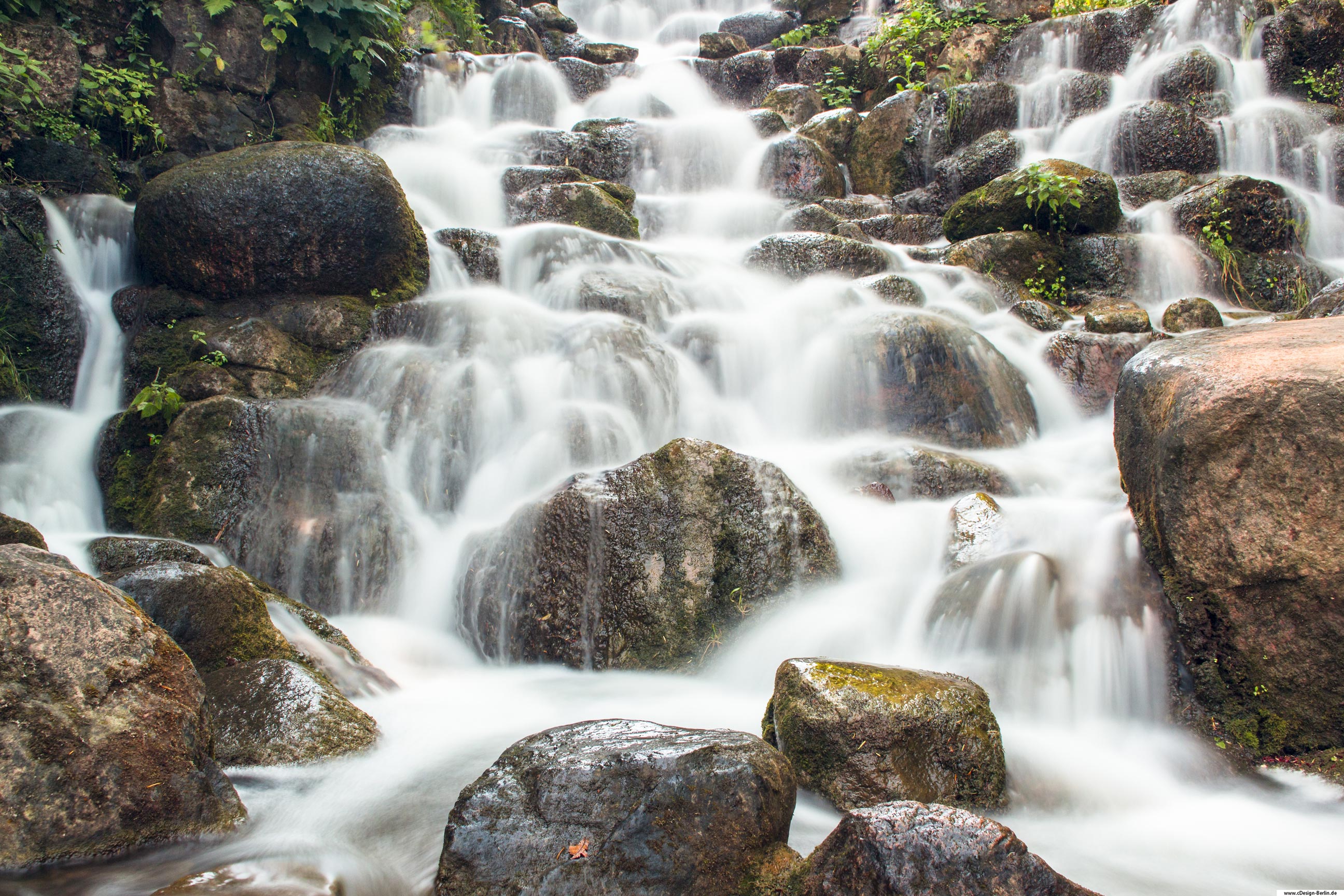Der Viktoriapark ist eine Grünanlage auf dem Kreuzberg, die höchste natürliche Erhebung der Berliner Innenstadt. Auf diesem Bild ist der Wasserfall des Viktoriaparks in einer Nahaufnahme mit Langzeitbelichtung mit Hilfe eines ND Filters zu sehen. Das Wasser fließt über eine Vielzahl von Steinen. Wer ein wenig Ruhe und ein Stück Oase sucht, wird in diesem Park nicht enttäuscht. Er ist neben dem Nationaldenkmal die bekannteste Attraktion des Viktoriaparks. Am Fuß des Wasserfalls wurde ein unregelmäßig begrenzter Teich angelegt. Bezirk: Berlin- Kreuzberg